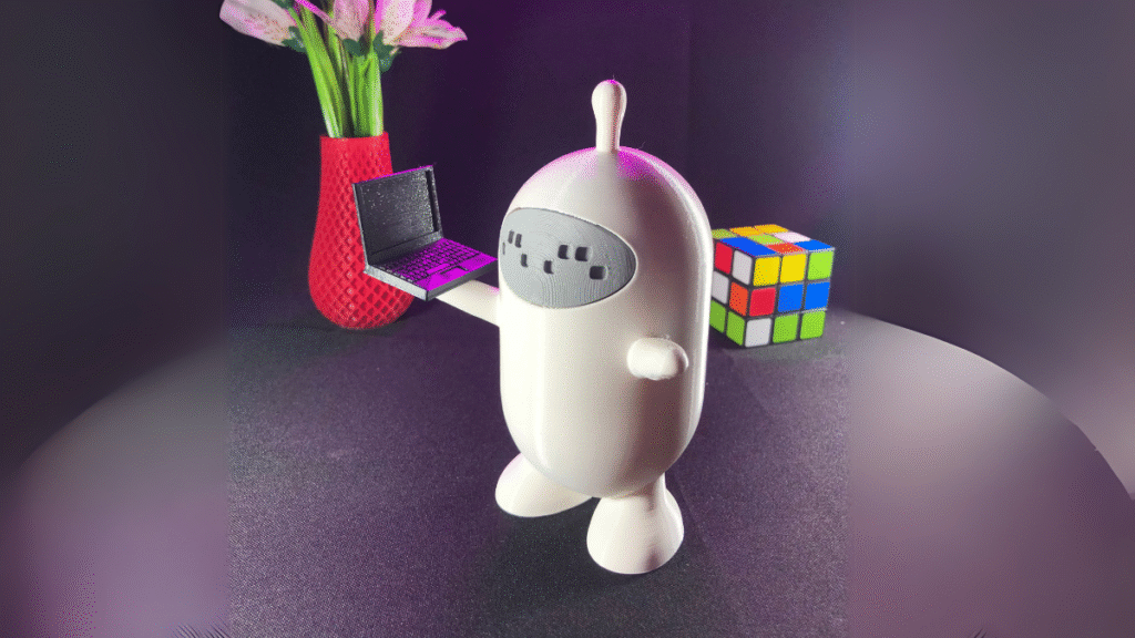 Gobotinho, mascote da GoBots impresso em 3D, segurando um mini notebook em mesa com vaso e cubo mágico