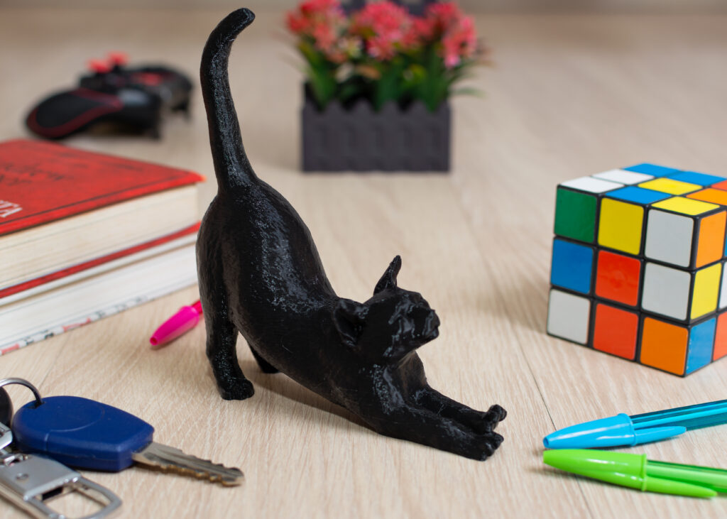 Gato em pose de alongamento impresso em 3D pela BR3D Soluções sobre mesa com canetas, cubo mágico e chaveiro